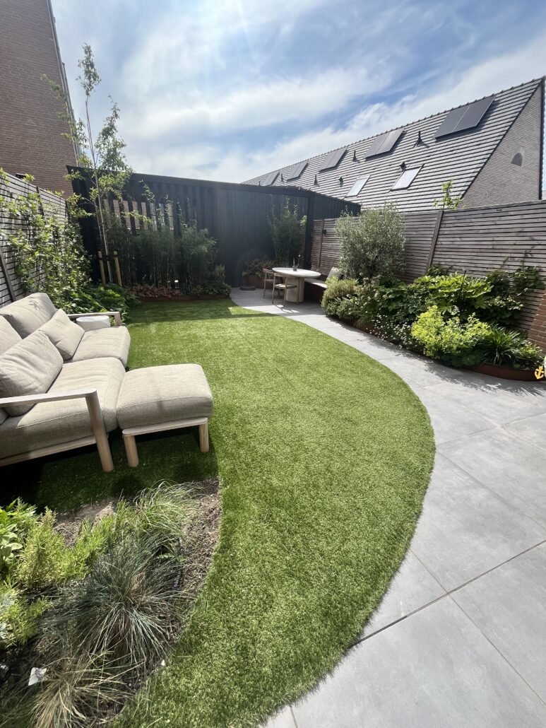 Foto van een aangelegde tuin met een gebogen gazon, grijze terrastegels, een loungebank en verhoogde borders van cortenstaal.
