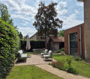 Overzicht van een loungeterras met groene meubels, een slingerend pad van Padvast, gazon en de houten uitbouw van de woning.