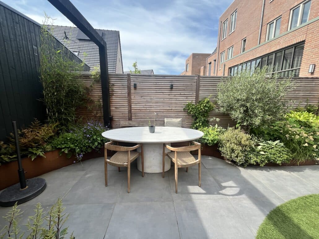 Rond terras van grijze tegels met een ovale eettafel en houten stoelen, geflankeerd door verhoogde borders van cortenstaal met groene beplanting.