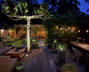 Verlichte achtertuin in de avond met lichtslingers, grondspots bij een grote leiboom, een houten vlonderterras met loungetuinmeubels en de overkapping op de achtergrond.