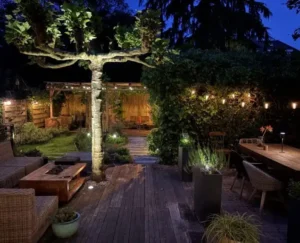 Verlichte achtertuin in de avond met lichtslingers, grondspots bij een grote leiboom, een houten vlonderterras met loungetuinmeubels en de overkapping op de achtergrond.