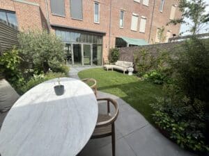 Terras met eettafel op de voorgrond en zicht op een rond gazon met loungebank, omgeven door een houten schutting en groene beplanting.