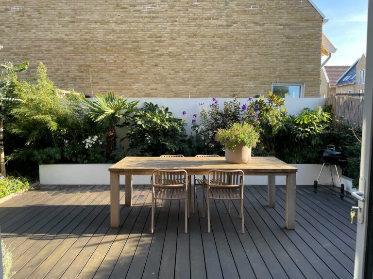Houten vlonderterras met een houten eettafel, witte verhoogde border en weelderige groene beplanting tegen een witte muur.